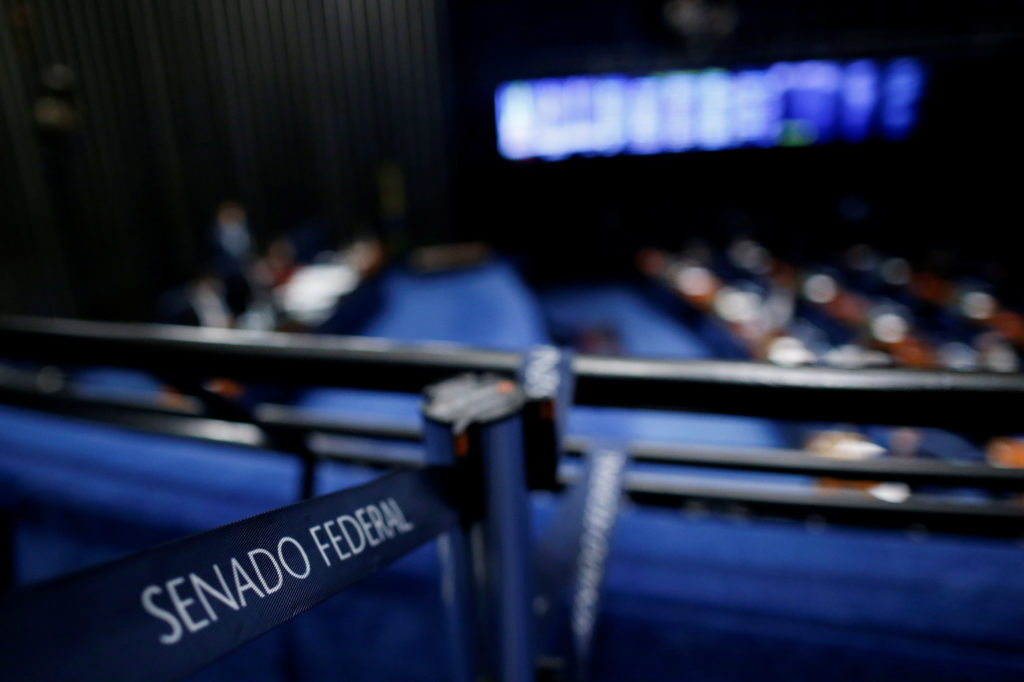 Faixa 'SENADO FEDERAL' em primeiro plano. Plenário com assentos azuis e parlamentares em sessão.
