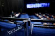 Faixa 'SENADO FEDERAL' em primeiro plano. Plenário com assentos azuis e parlamentares em sessão.