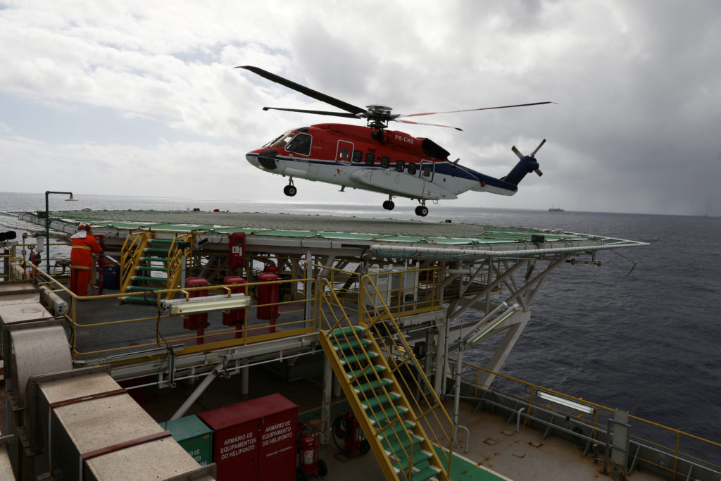 Helicóptero vermelho e branco pairando sobre heliponto de plataforma offshore.