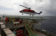 Helicóptero vermelho e branco pairando sobre heliponto de plataforma offshore.