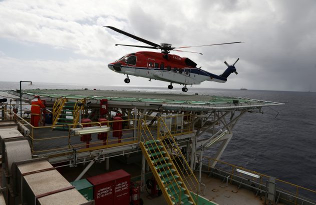 Helicóptero vermelho e branco pairando sobre heliponto de plataforma offshore.