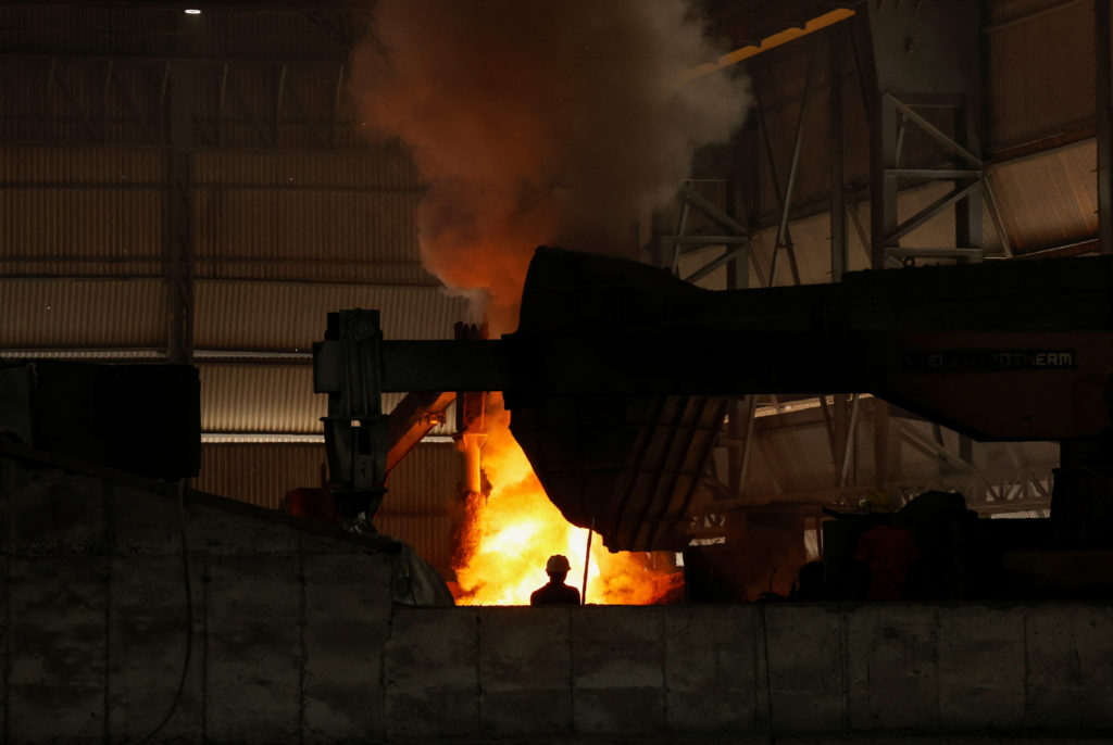 Fundição: operário em silhueta observa metal incandescente e fumaça intensa em ambiente industrial.