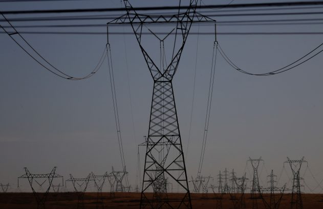 Silhuetas de muitas torres de alta tensão e suas linhas sobre um campo sob céu claro-escuro, com uma torre central.