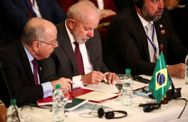 Lula escreve em documentos numa mesa de reunião com outros dois homens e uma bandeira do Brasil.