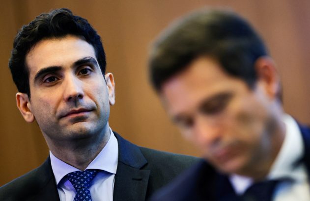 Homem de terno escuro e gravata azul olha atento e pensativo. Outro homem desfocado ao lado.