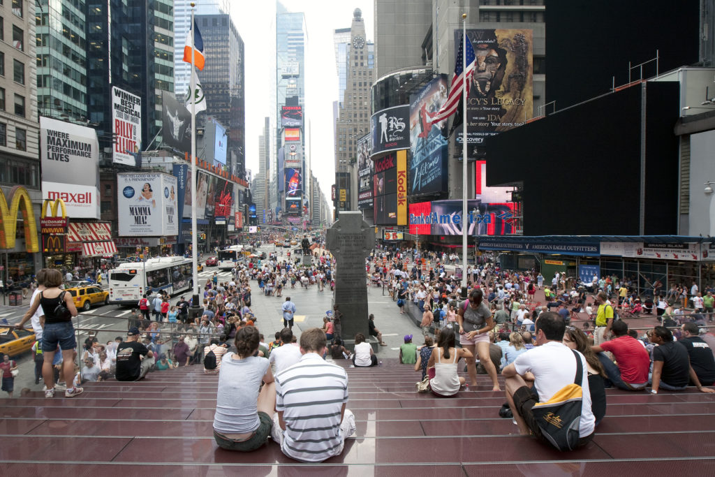 Times Square movimentada com multidões, painéis publicitários e pessoas sentadas em degraus.