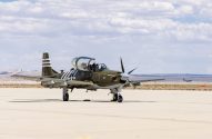 Avião militar verde-oliva com hélice, parado em pátio de aeroporto.