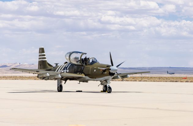 Avião militar verde-oliva com hélice, parado em pátio de aeroporto.