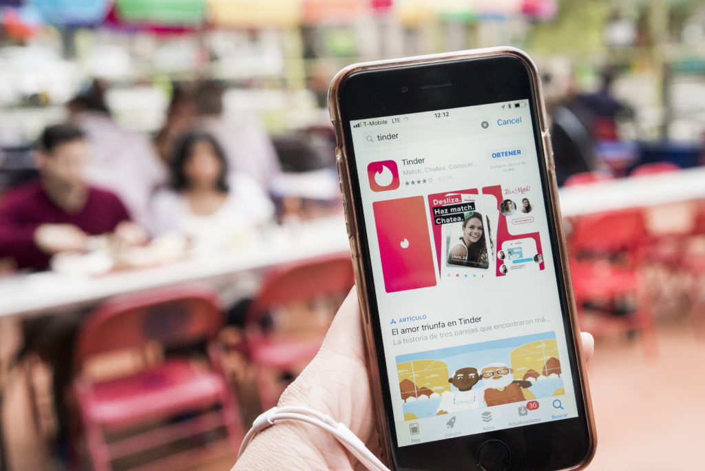 Pessoa usando celular mostra app Tinder na App Store, com fundo borrado de pessoas.