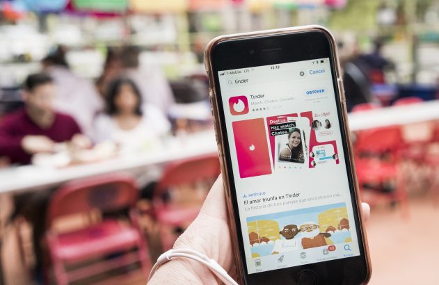 Pessoa usando celular mostra app Tinder na App Store, com fundo borrado de pessoas.
