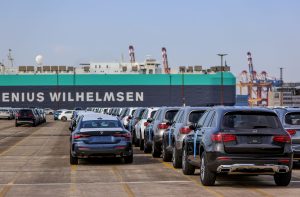 Fileiras de carros novos em pátio de porto, prontos para embarque em grande navio cargueiro.