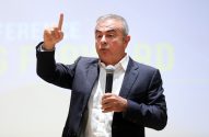 Carlos Ghosn, em terno, segura microfone e aponta o dedo indicador para cima durante uma apresentação.