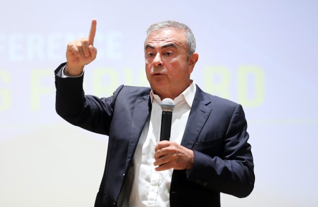 Carlos Ghosn, em terno, segura microfone e aponta o dedo indicador para cima durante uma apresentação.