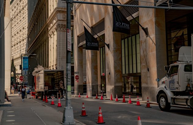 Rua urbana com prédios altos. Caminhões (um soltando vapor), cones laranja na via e pessoas caminhando na calçada.