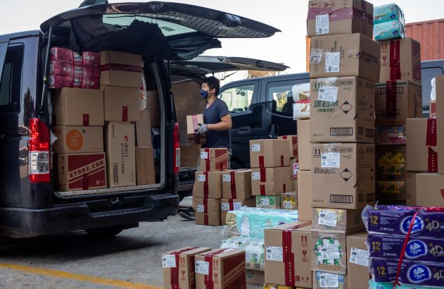 Homem de máscara descarrega van com caixas de produtos de limpeza, máscaras e itens diversos, e pilhas de caixas.