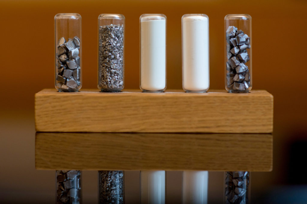 Cinco frascos exibem diferentes formas de um material: pedaços e granulado metálico, e pó branco, sobre madeira.
