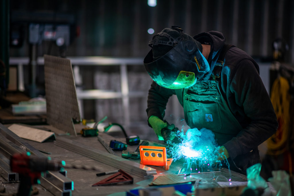 Soldador de capacete emitindo faíscas azuis brilhantes ao soldar metal em uma oficina.