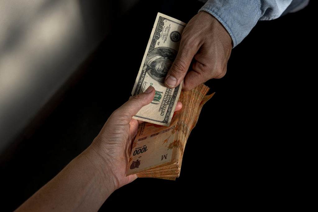 Duas mãos trocam dinheiro. Uma entrega uma nota de dólar, a outra segura notas de reais.