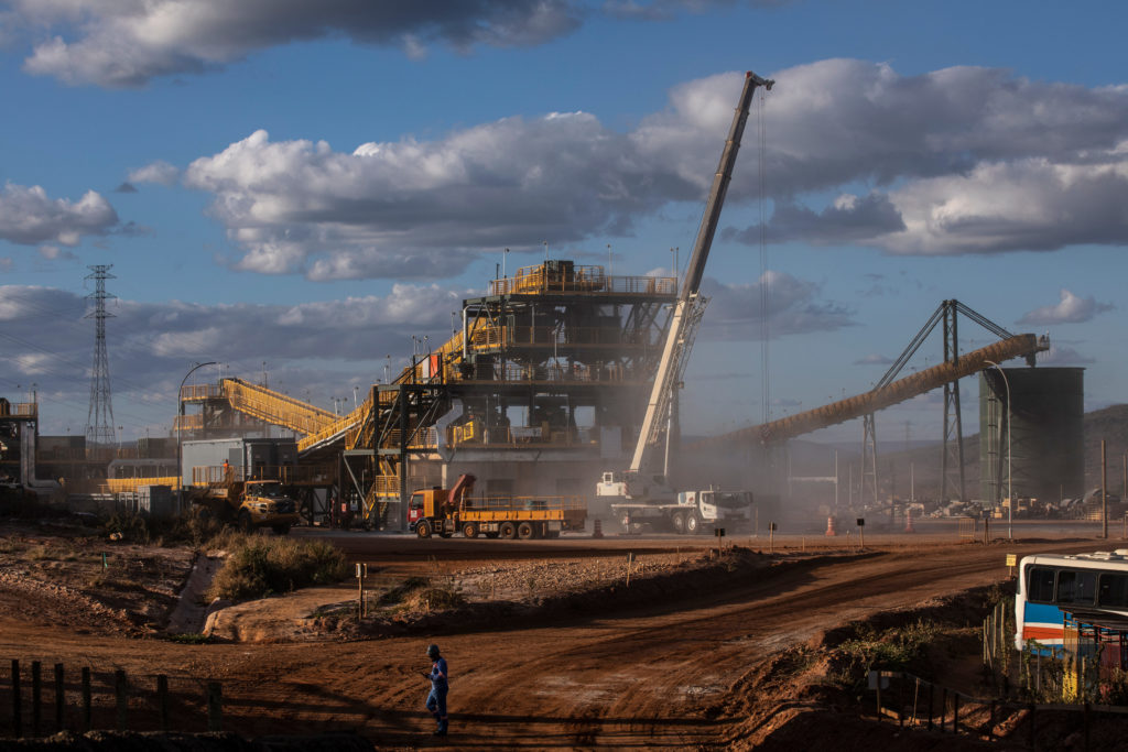 Operação industrial de mineração com estruturas amarelas, esteiras transportadoras, guindaste, caminhões, poeira e céu azul.