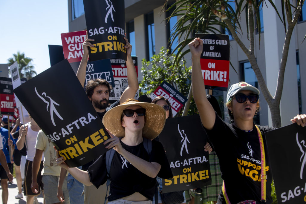 Manifestantes SAG-AFTRA em greve marcham com cartazes e punhos erguidos sob o sol.