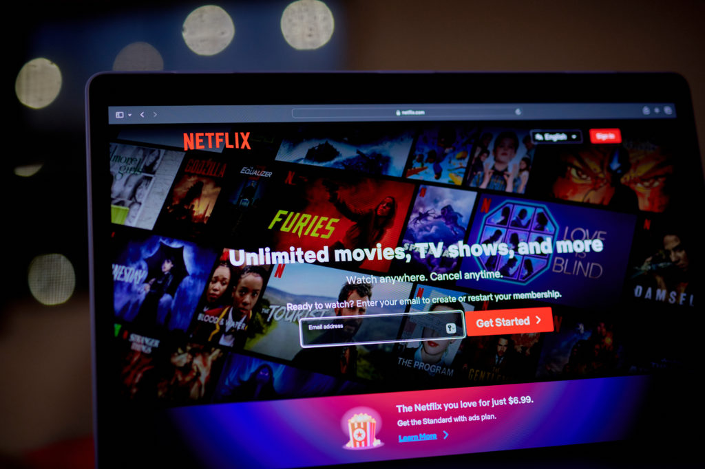 Tela de notebook mostrando a página inicial da Netflix com títulos de conteúdo e campo de email para inscrição.