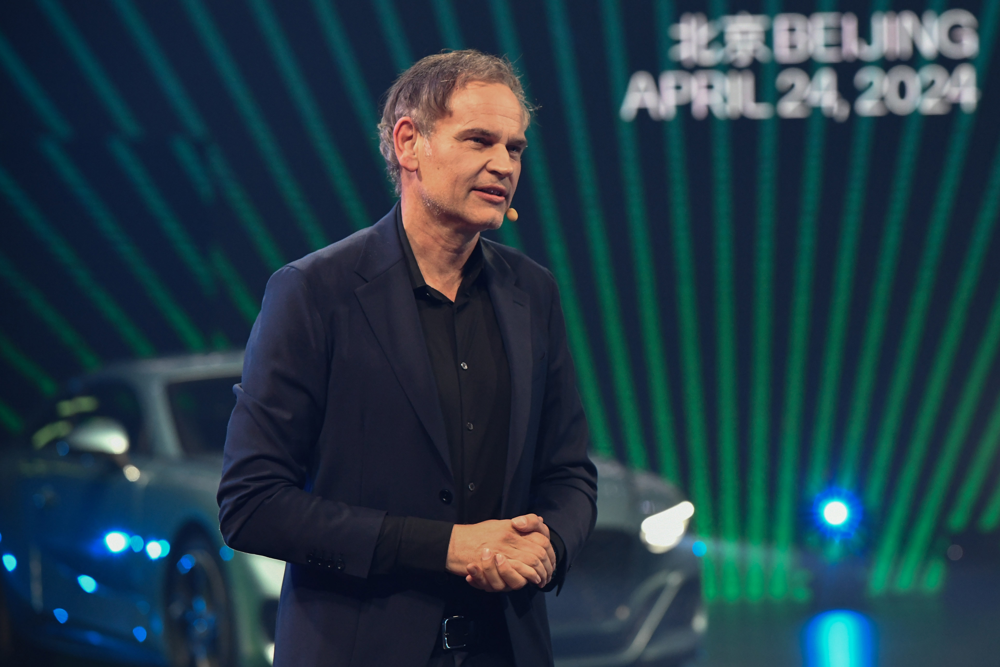 Homem discursa em palco com carros futuristas no fundo. Texto desfocado: 'BEIJING APRIL 24, 2024'.