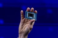 Mão segura um chip eletrônico retangular contra um fundo azul desfocado.