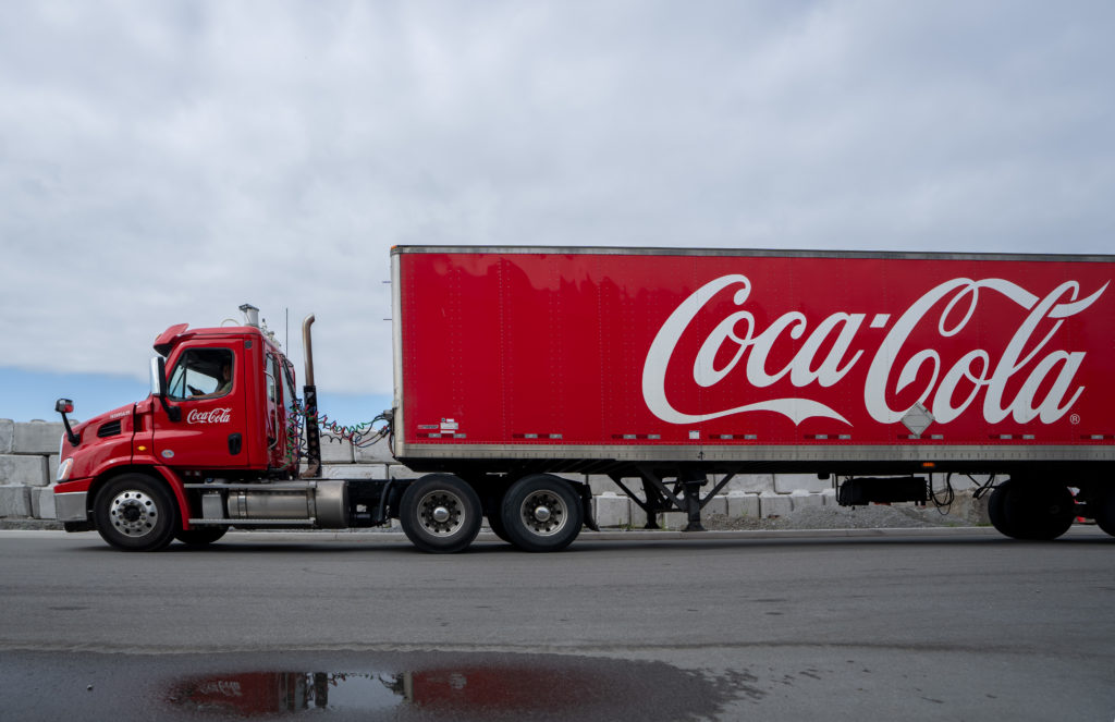 Caminhão vermelho da Coca-Cola com logo branco no reboque, em via asfaltada.