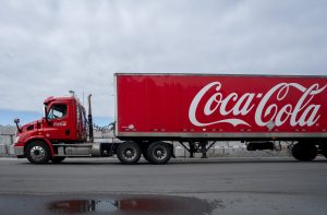 Caminhão vermelho da Coca-Cola com logo branco no reboque, em via asfaltada.