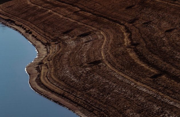 Vista aérea de reservatório com nível de água baixo, revelando margem seca em camadas de terra escura.