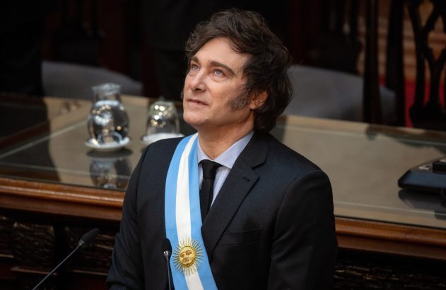 Javier Milei, presidente da Argentina, com faixa presidencial azul e branca, olha para o alto em evento.