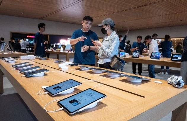 Funcionário auxilia cliente com celular em loja. Mesas de madeira com iPads e celulares expostos para interação.