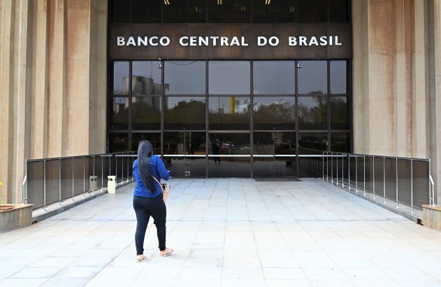 Pessoa de costas caminha em direção ao Banco Central do Brasil, prédio com fachada de vidro.
