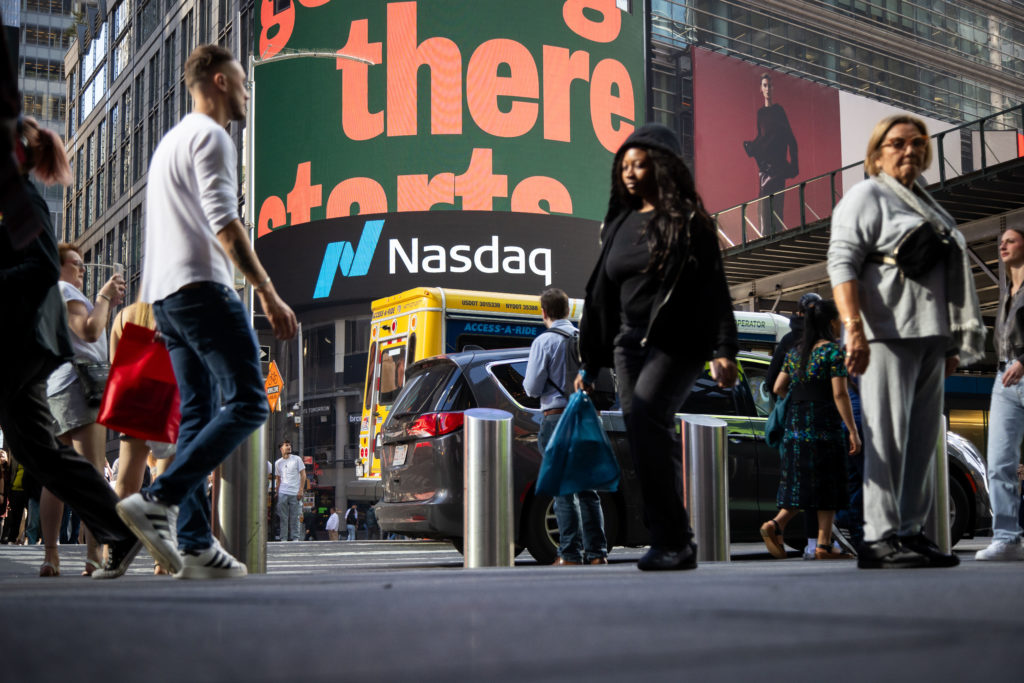 Vista em ângulo baixo de pedestres caminhando em rua movimentada com o letreiro gigante da Nasdaq ao fundo.