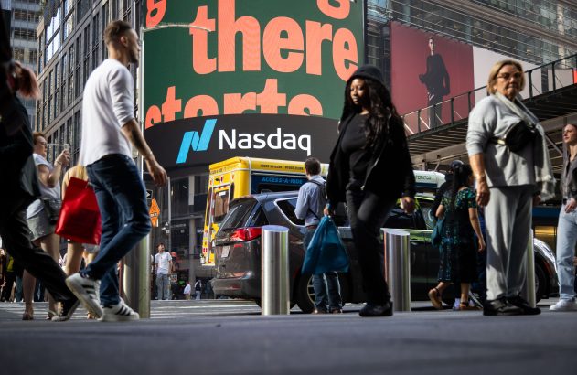 Vista em ângulo baixo de pedestres caminhando em rua movimentada com o letreiro gigante da Nasdaq ao fundo.