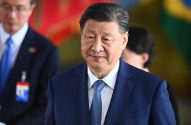 Xi Jinping, de terno azul, olha levemente para a direita com um sorriso sutil. Pessoas e cores desfocadas ao fundo.