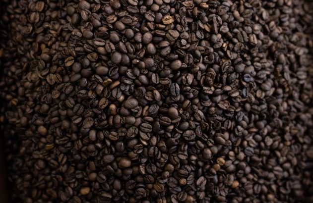 Grande pilha de grãos de café torrado escuro, preenchendo toda a imagem. Detalhe da textura irregular e tons variados.