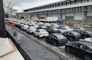 Filas de sedans zero-quilômetro brancos e pretos em cais. Trabalhadores preparam veículos para embarque ou desembarque.
