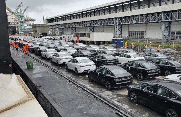 Filas de sedans zero-quilômetro brancos e pretos em cais. Trabalhadores preparam veículos para embarque ou desembarque.