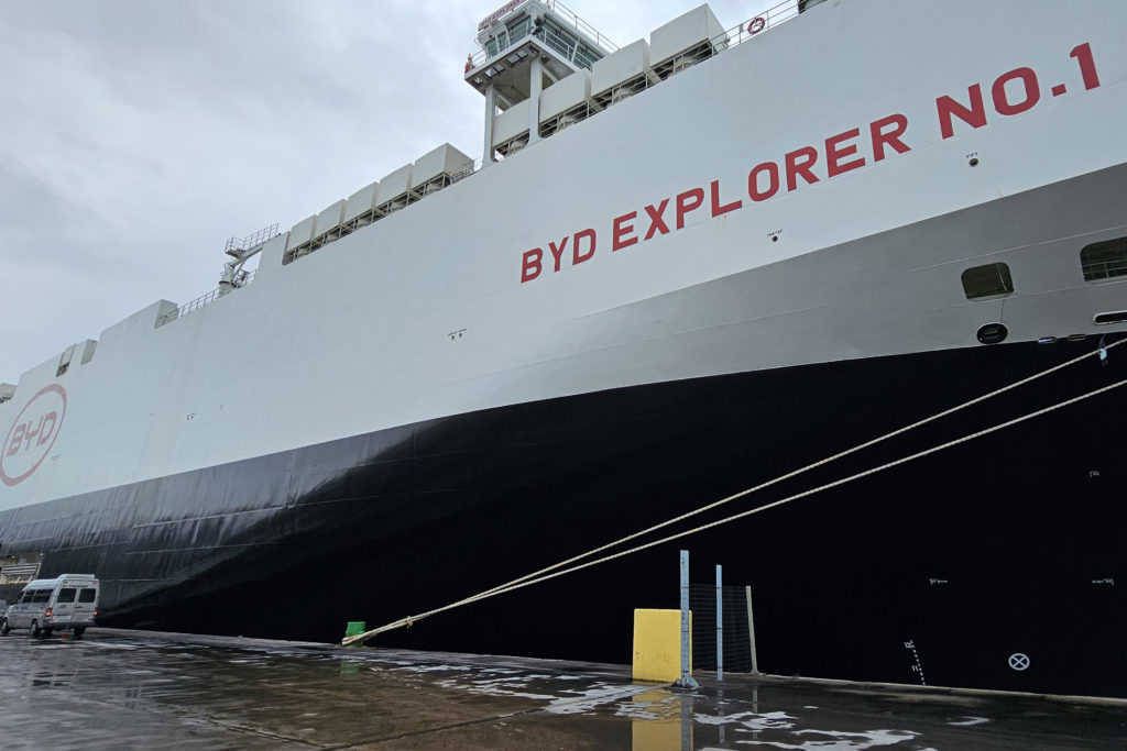 Navio BYD EXPLORER NO. 1, com logo BYD, atracado em porto com chão molhado e vans próximas.
