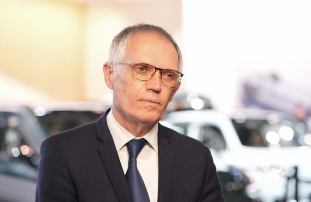 Homem de terno e óculos olhando para o lado. Fundo desfocado com carros e luzes.