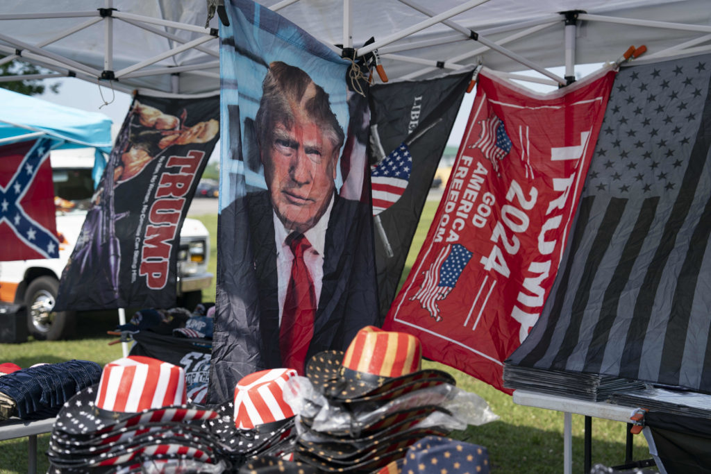 Banca sob tenda com bandeiras e chapéus. Destaque para imagens de Trump, bandeira confederada e 'TRUMP 2024'.