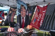Banca sob tenda com bandeiras e chapéus. Destaque para imagens de Trump, bandeira confederada e 'TRUMP 2024'.