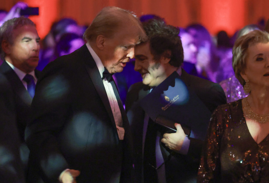 Donald Trump, em smoking, inclinado para falar com homem de cabelo escuro, que sorri e segura uma pasta azul.