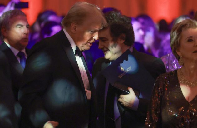 Donald Trump, em smoking, inclinado para falar com homem de cabelo escuro, que sorri e segura uma pasta azul.