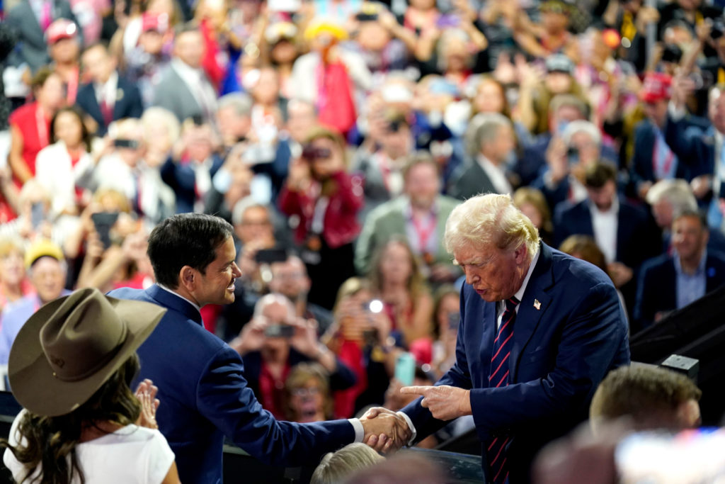 Donald Trump aperta a mão de um homem, com multidão ao fundo.
