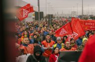 Manifestação: trabalhadores carregam bandeiras vermelhas com logo do sindicato IG Metall, sob céu cinzento.