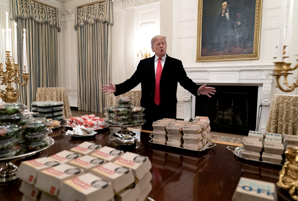 Donald Trump com braços abertos atrás de mesa com grandes pilhas de dinheiro em sala.