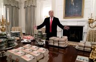 Donald Trump com braços abertos atrás de mesa com grandes pilhas de dinheiro em sala.