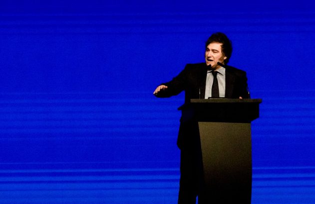 Homem em terno discursa em pódio, gesticulando contra fundo azul vibrante.